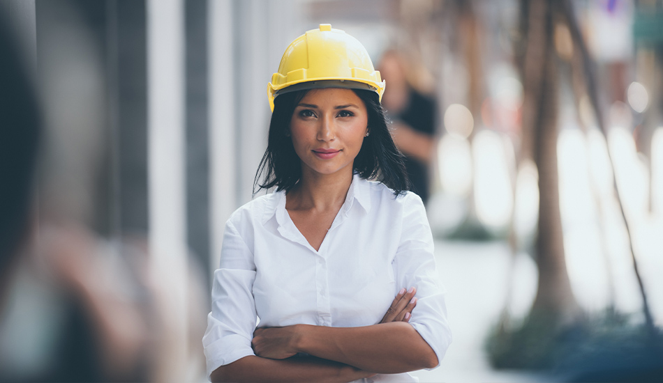 Frau steht vor einer Baustelle mit einem Helm auf dem Kopf und verschränkten Armen